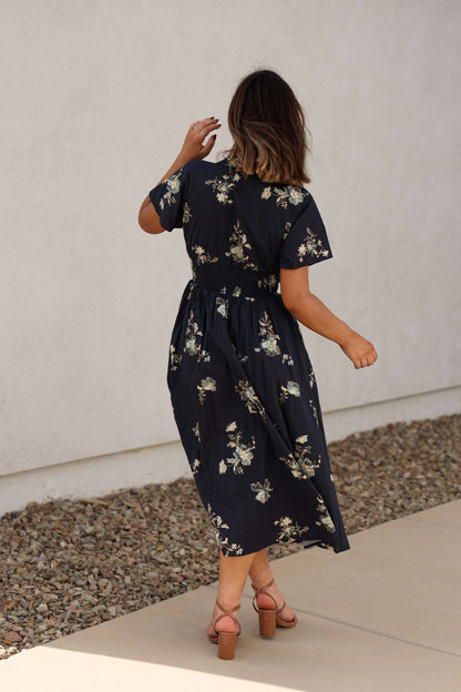 Woman in a Pumpkin Spice Black Floral Zip Midi Dress and sandals walks on a sidewalk, light wall behind her.
