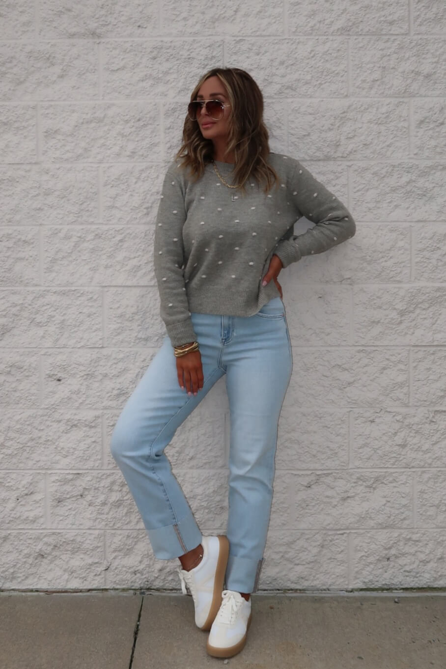 A woman in sunglasses, light blue jeans, and white sneakers wears a Ramona Sage Polka Dot Ribbed Sweater by a white brick wall.