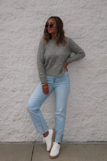 A woman in sunglasses, light blue jeans, and white sneakers wears a Ramona Sage Polka Dot Ribbed Sweater by a white brick wall.