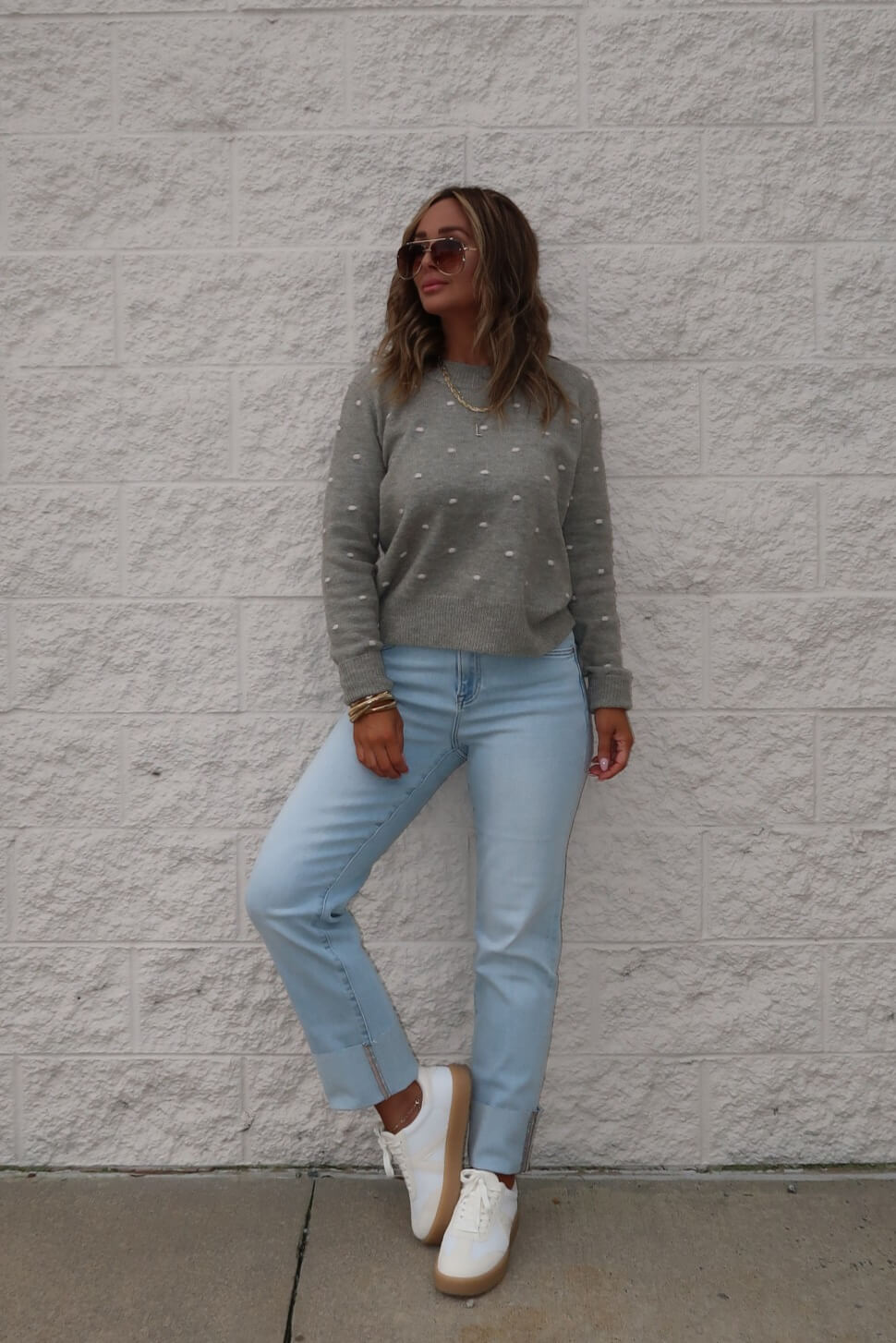 Woman in sunglasses, light blue jeans, and white sneakers wears a Ramona Sage Polka Dot Ribbed Sweater by a light brick wall.