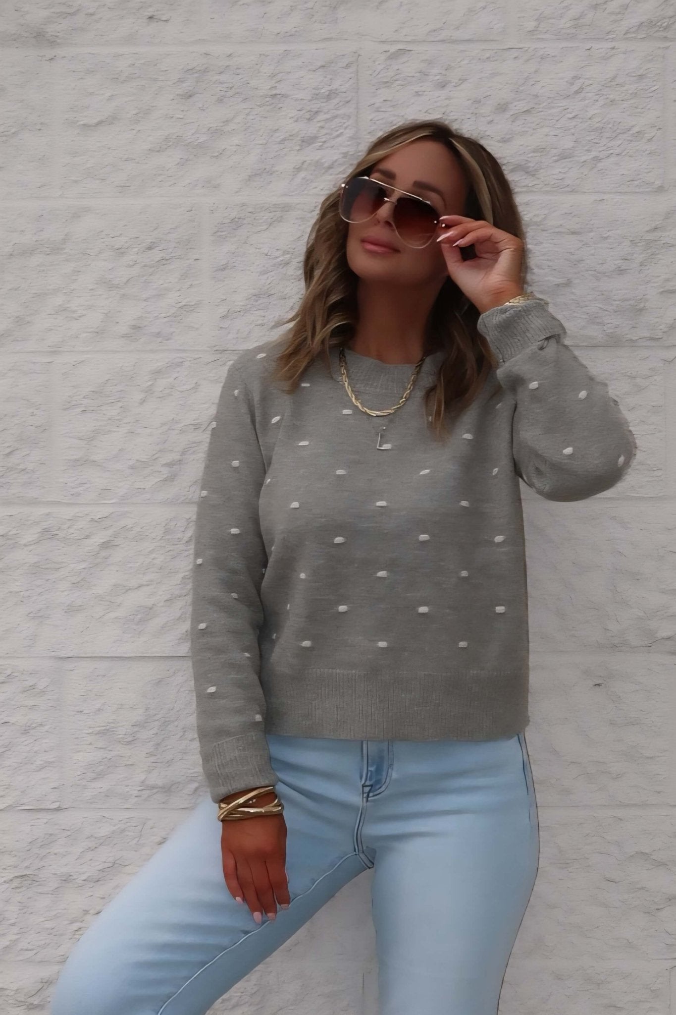 A woman in the Ramona Sage Polka Dot Ribbed Sweater and light jeans poses, accessorized with sunglasses and layered necklaces.