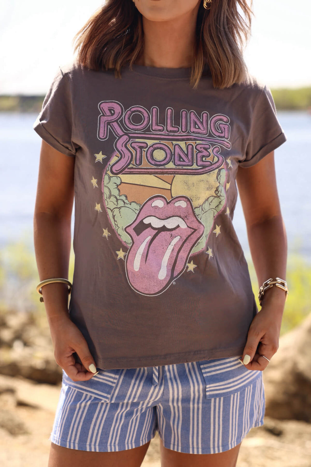 Woman wears the Recycled Karma Grey Rolling Stones 1978 Graphic Tee by the water, hands resting on her shorts.