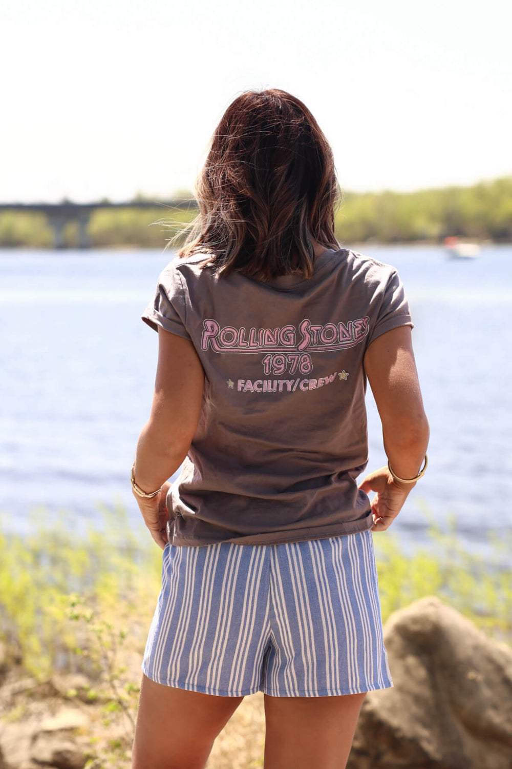 A woman stands by a lake in a Recycled Karma Grey Rolling Stones 1978 Graphic Tee, soaking up the sunny day.