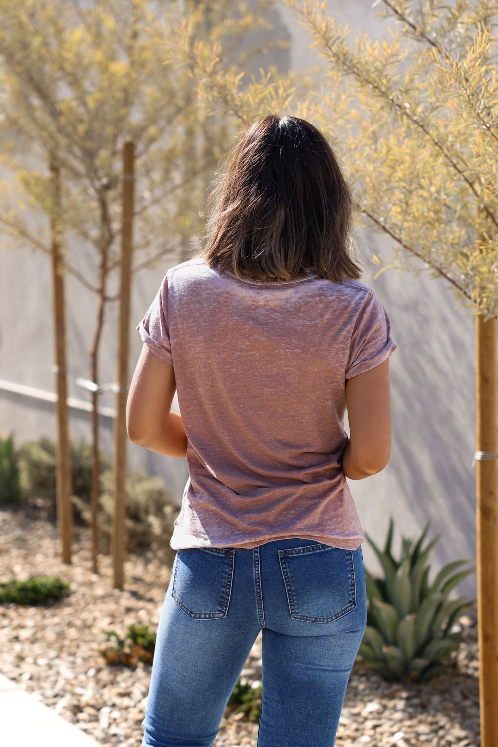A woman with shoulder-length hair stands outdoors in the Recycled Karma Mocha Miller Graphic Tee and blue jeans near young trees.