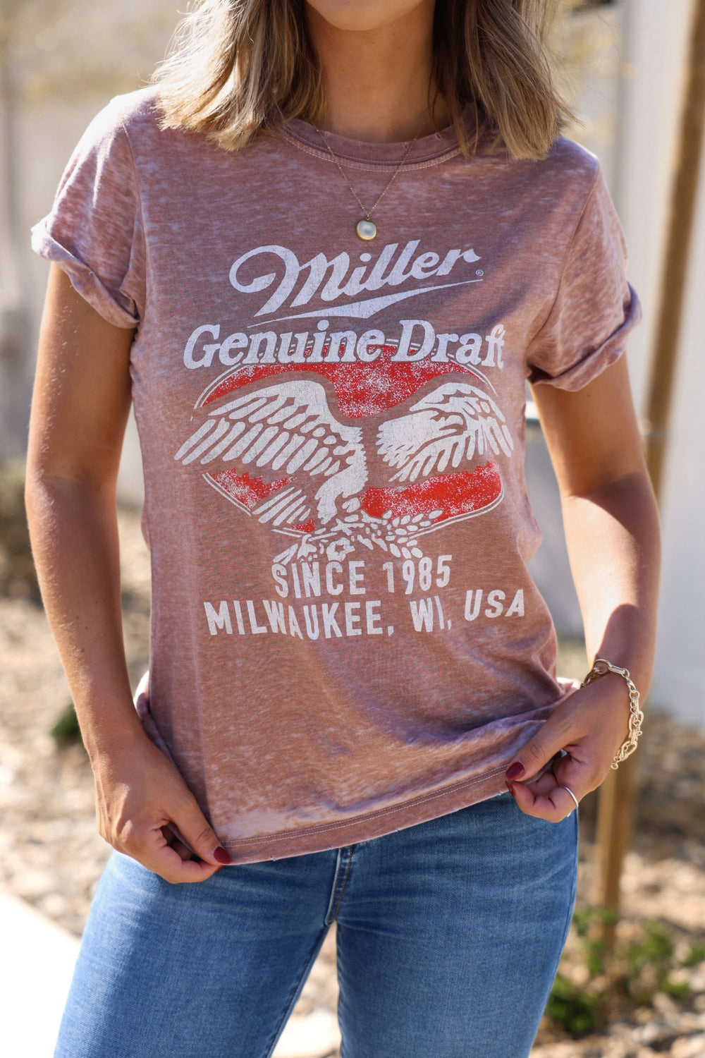 Woman wearing the Recycled Karma Mocha Miller Graphic Tee, paired with jeans, standing outdoors—torso and arms shown.