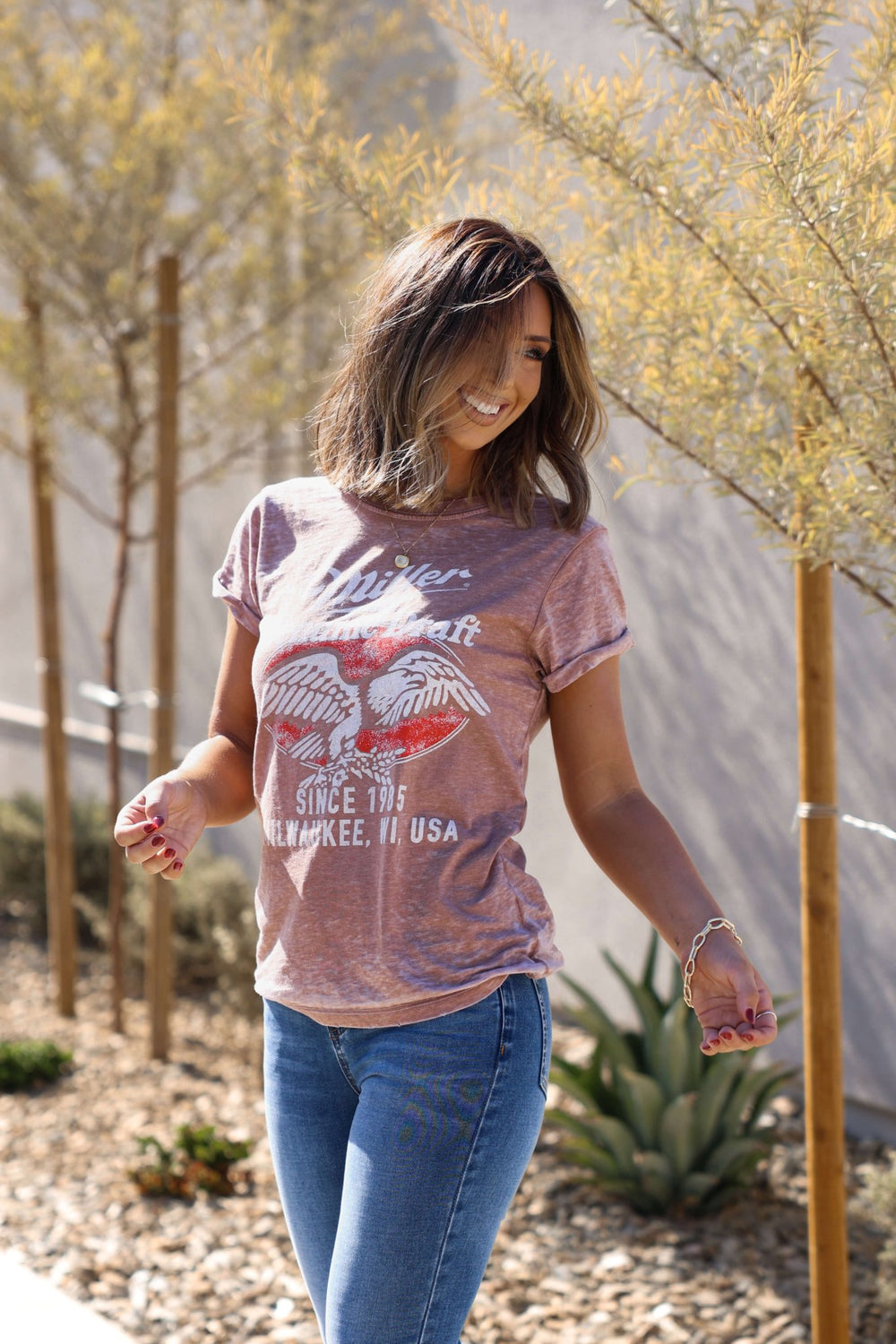 Woman smiles outdoors in the Recycled Karma Mocha Miller Graphic Tee, surrounded by plants and trees on a sunny day.