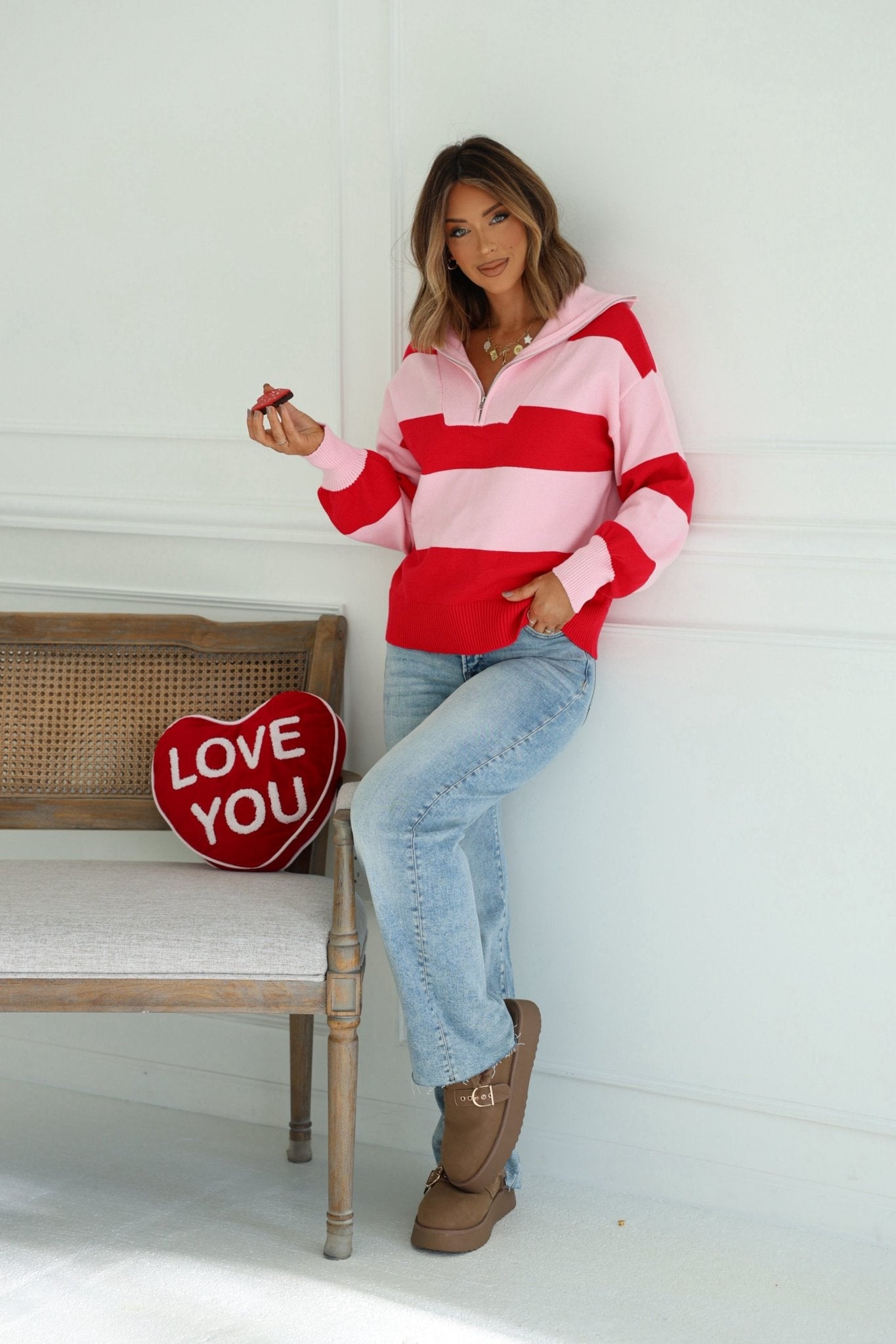 Woman in a Red and Pink Stripe Quarter Zip Sweater with balloon sleeves holds chocolate by a bench with a red LOVE YOU heart pillow.