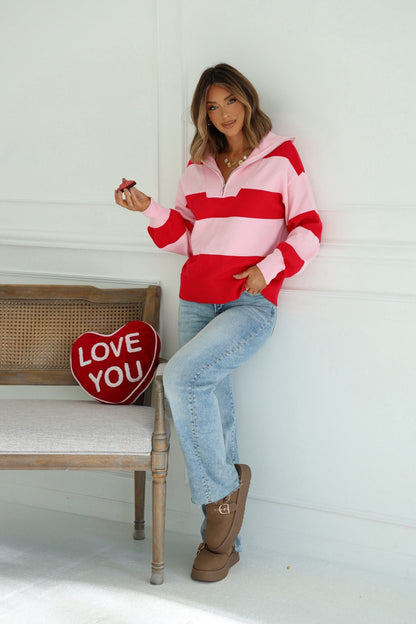 Woman in a Red and Pink Stripe Quarter Zip Sweater with balloon sleeves holds chocolate by a bench with a red LOVE YOU heart pillow.