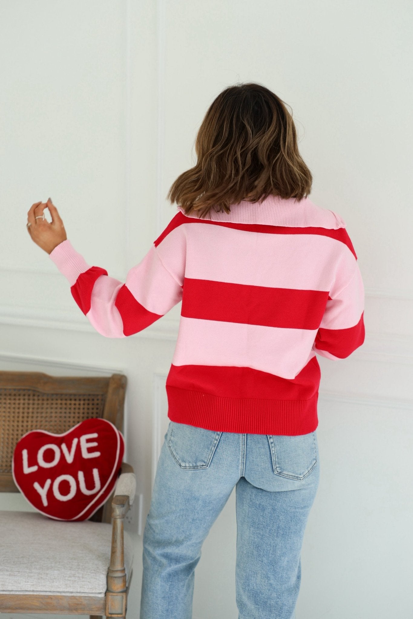 A woman in a Red and Pink Stripe Quarter Zip Sweater stands facing away near a chair with a red heart LOVE YOU pillow.