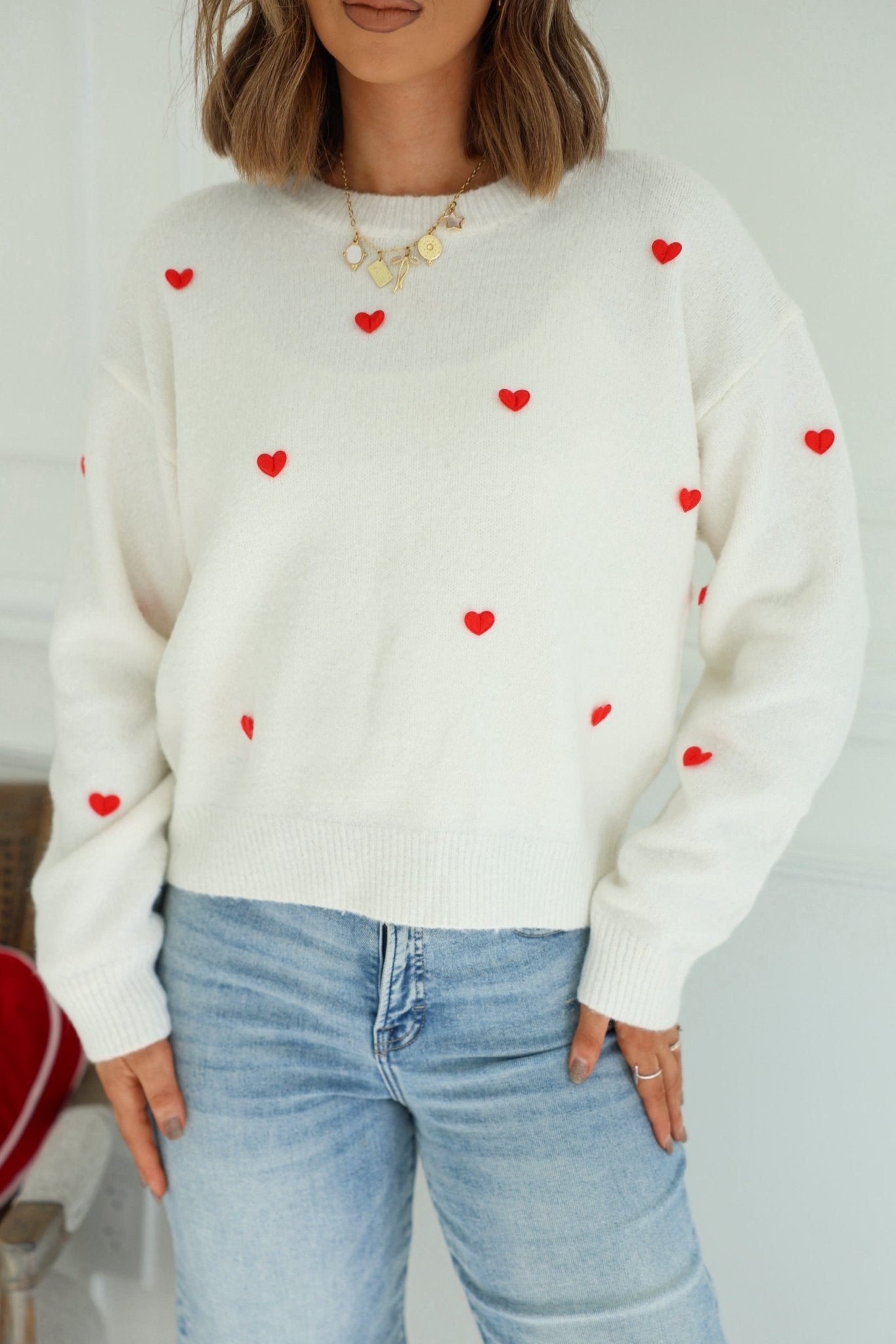 Woman in a Red and White 3D Heart Sweater, light blue jeans, and gold necklace standing indoors.