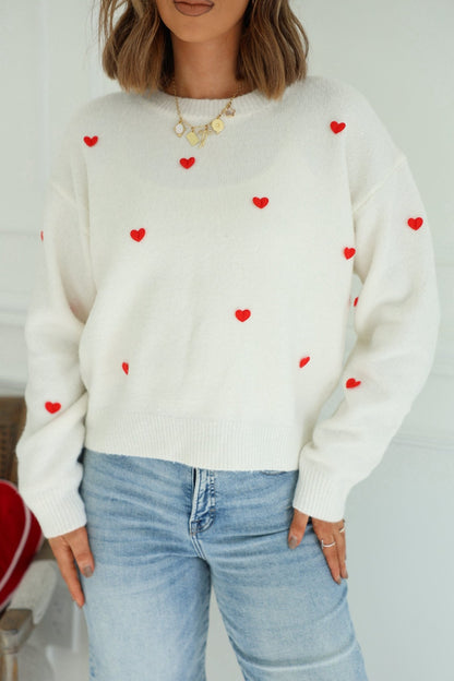 Woman in a Red and White 3D Heart Sweater, light blue jeans, and gold necklace standing indoors.