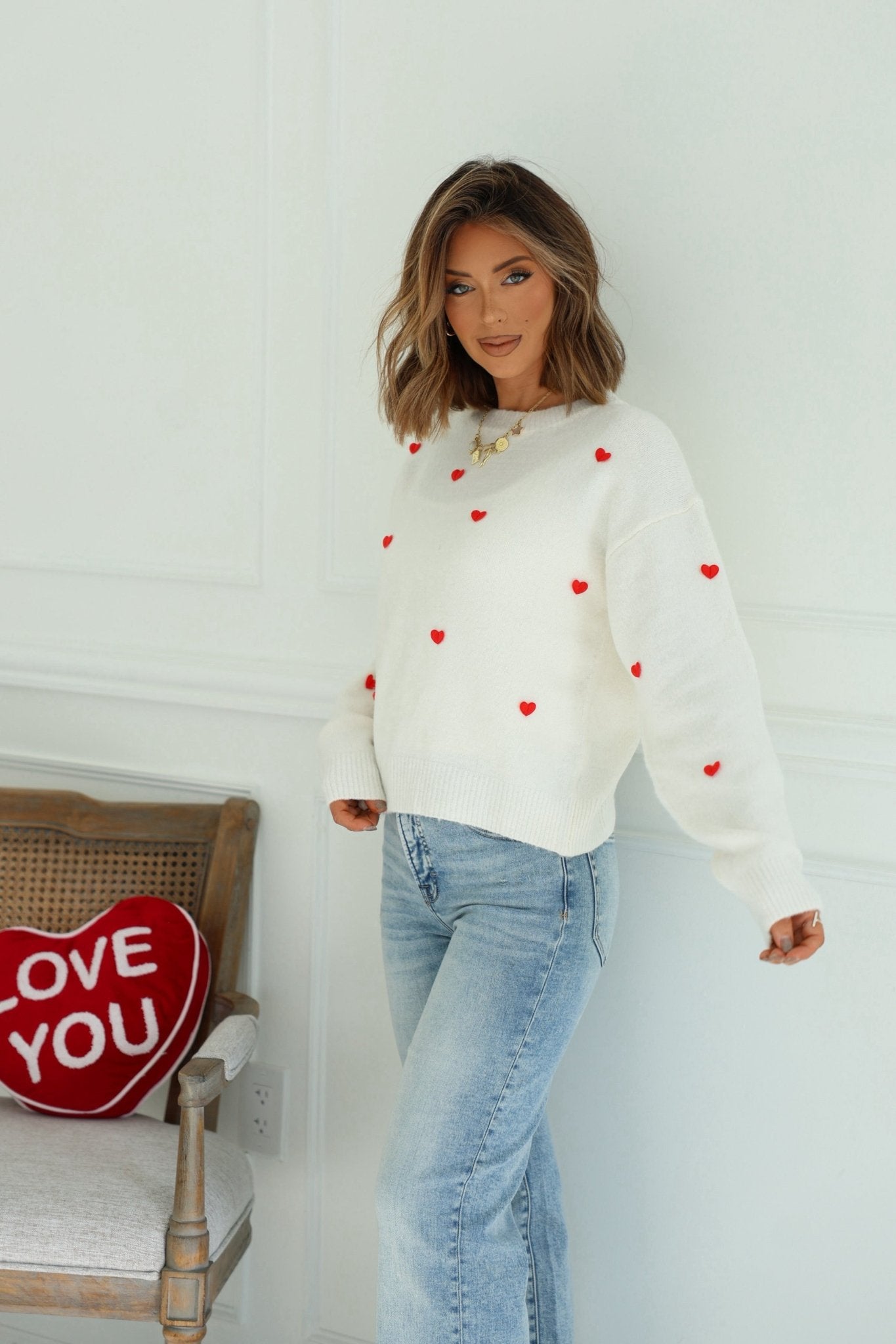 Woman wears a Red and White 3D Heart Sweater with blue jeans, standing by a chair with a red LOVE YOU pillow.
