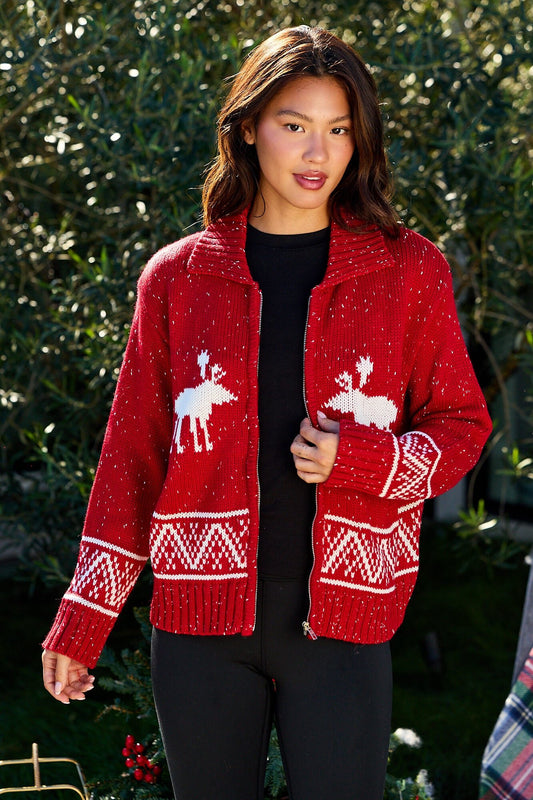 A woman wears a Red and White Aztec Zip Sweater Cardigan outdoors in front of green foliage.