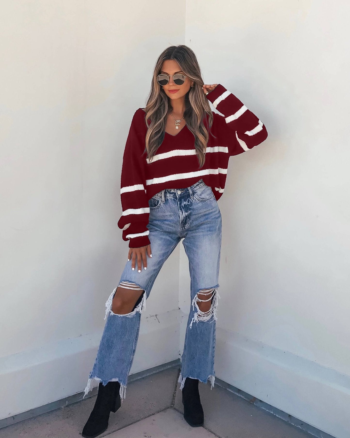 Woman in a chic fall look wears a Red and White Striped V Neck Sweater with ripped jeans, black boots, and sunglasses.