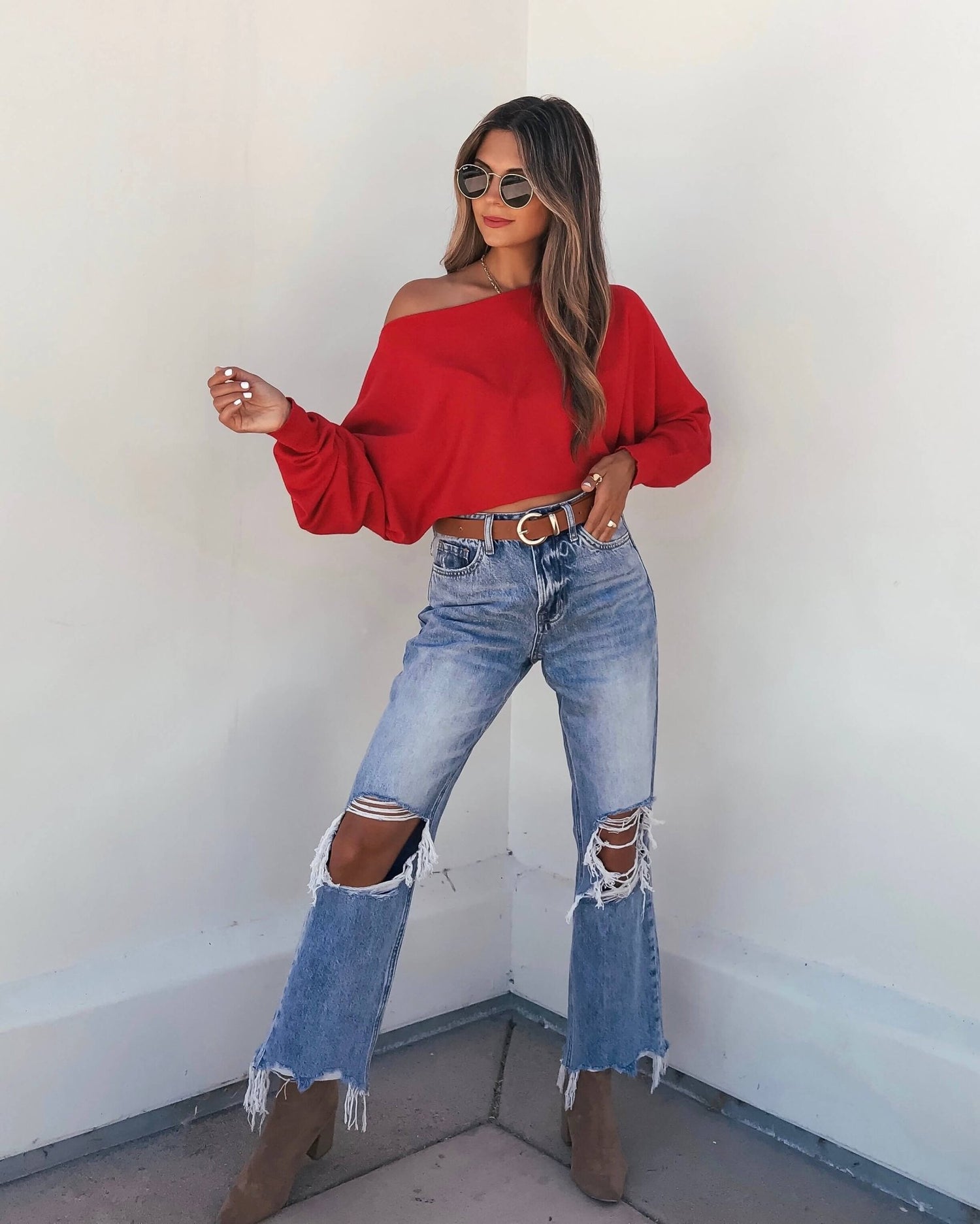 Woman in sunglasses wears a Red Boatneck Cashmere Sweater, ripped jeans, brown belt, and ankle boots against a white wall.