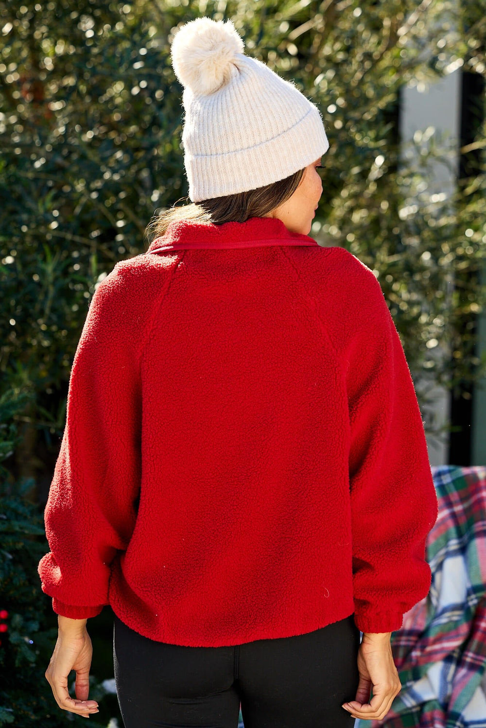 A person in a Red Button Down Sherpa Jacket and white knit beanie stands outdoors with greenery behind them.