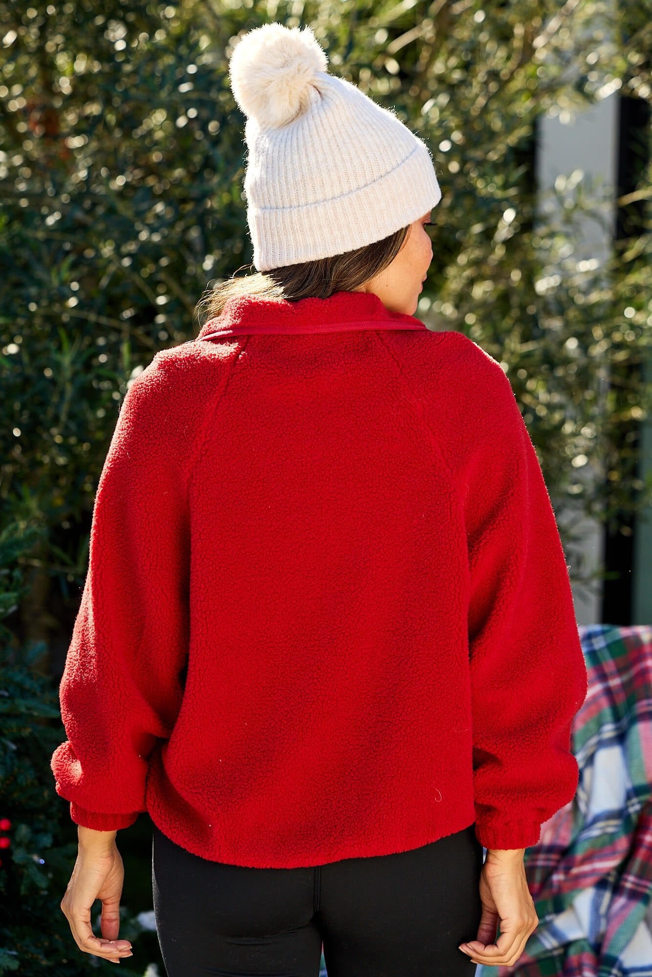 A person in a Red Button Down Sherpa Jacket and white knit beanie stands outdoors with greenery behind them.
