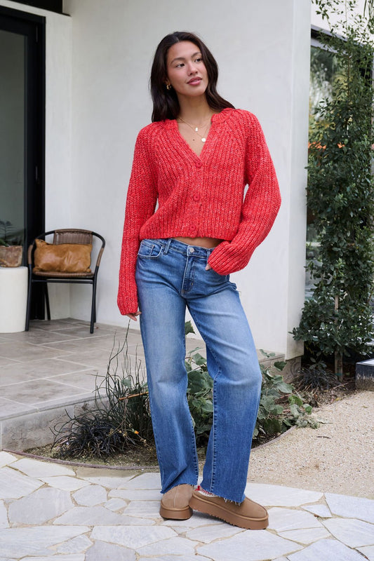 Woman outdoors in a Red Confetti Knit Buttoned Cardigan - FINAL SALE, blue jeans, and tan shoes, with plants and a chair behind her.