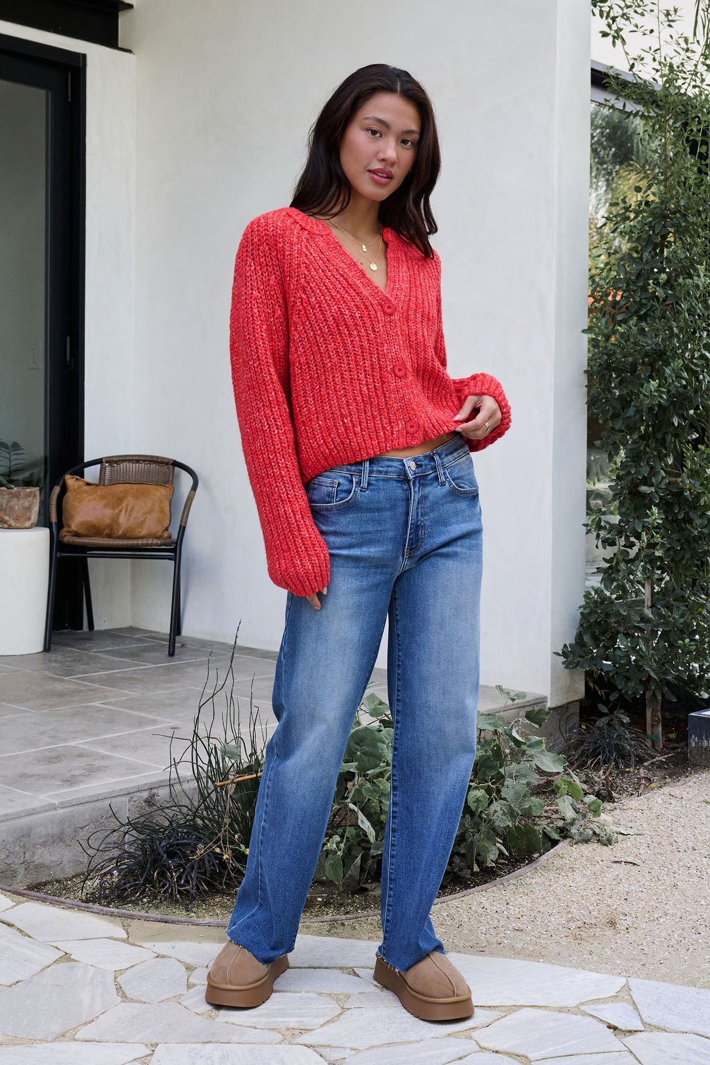 Woman outdoors in a Red Confetti Knit Buttoned Cardigan, blue jeans, and tan shoes with greenery and a modern house behind her.