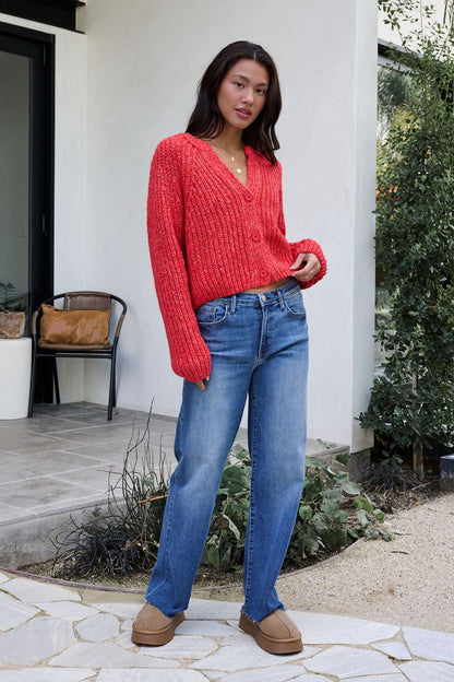 Woman outdoors in a Red Confetti Knit Buttoned Cardigan, blue jeans, and tan shoes with greenery and a modern house behind her.