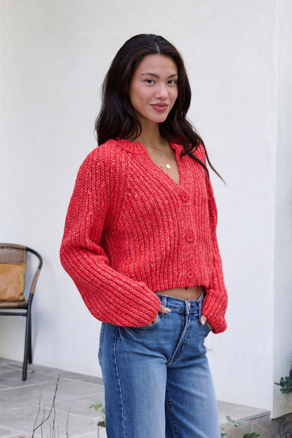 A woman in a Red Confetti Knit Buttoned Cardigan and jeans stands outside, smiling with one hand in her pocket.