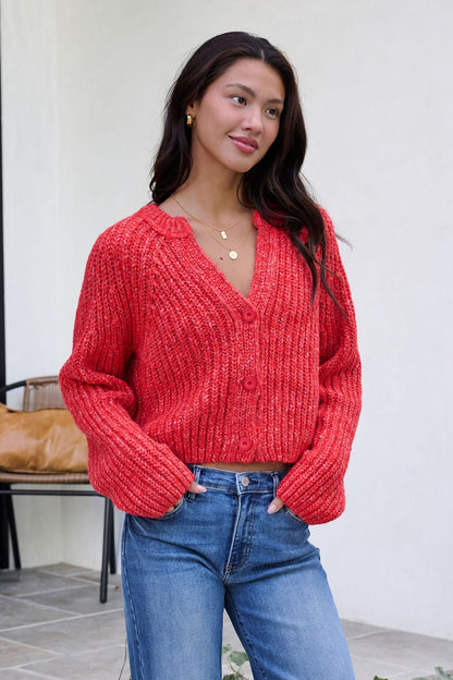 Woman in a Red Confetti Knit Buttoned Cardigan and blue jeans stands outdoors, hands in pockets, smiling softly to the side.