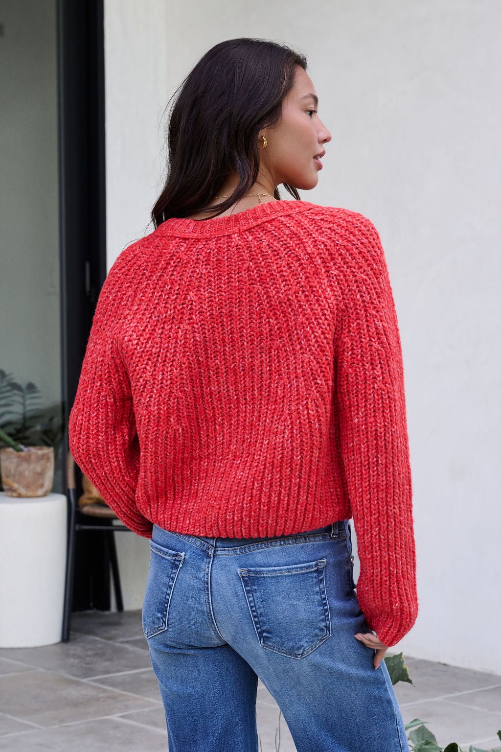 A woman in a Red Confetti Knit Buttoned Cardigan and blue jeans stands outside, facing away from the camera.