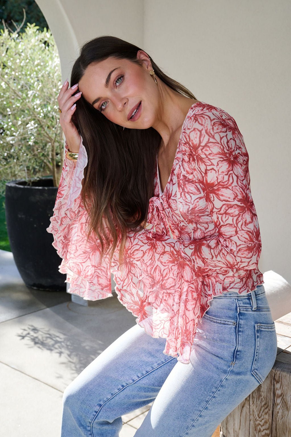 Woman sitting on a bench in a red Floral Print Bell Sleeve Mesh Top and jeans, touching her hair, with plants in the background.