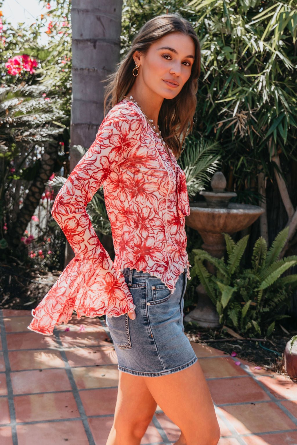 Woman outdoors in Red Floral Print Bell Sleeve Mesh Top - FINAL SALE, denim mini skirt, on a tiled patio with greenery.