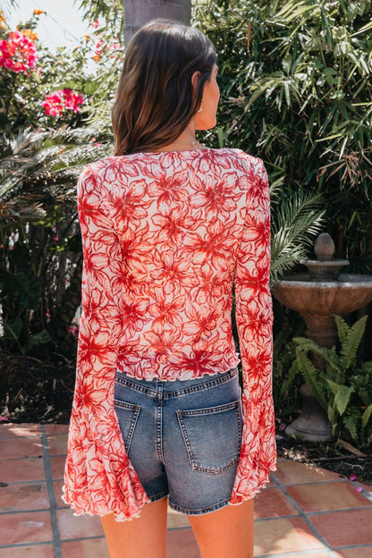 A woman with wavy brown hair wears the Red Floral Print Bell Sleeve Mesh Top, paired with denim shorts, standing outdoors by green plants.