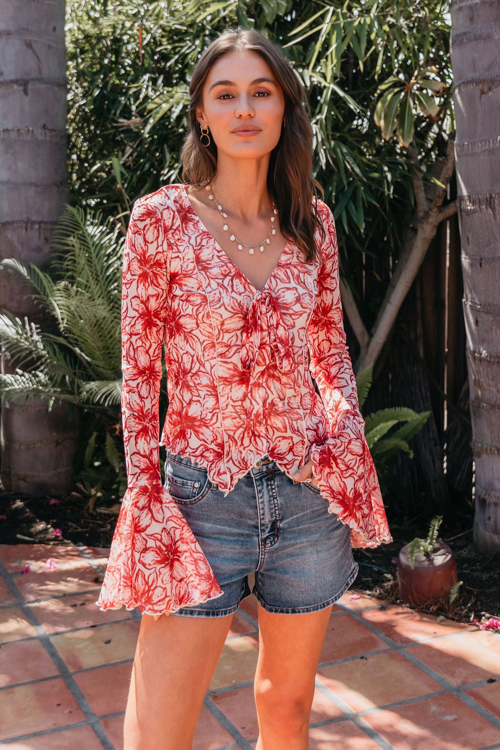 Woman outdoors in a Red Floral Print Bell Sleeve Mesh Top - FINAL SALE, paired with denim shorts among greenery and potted plants.