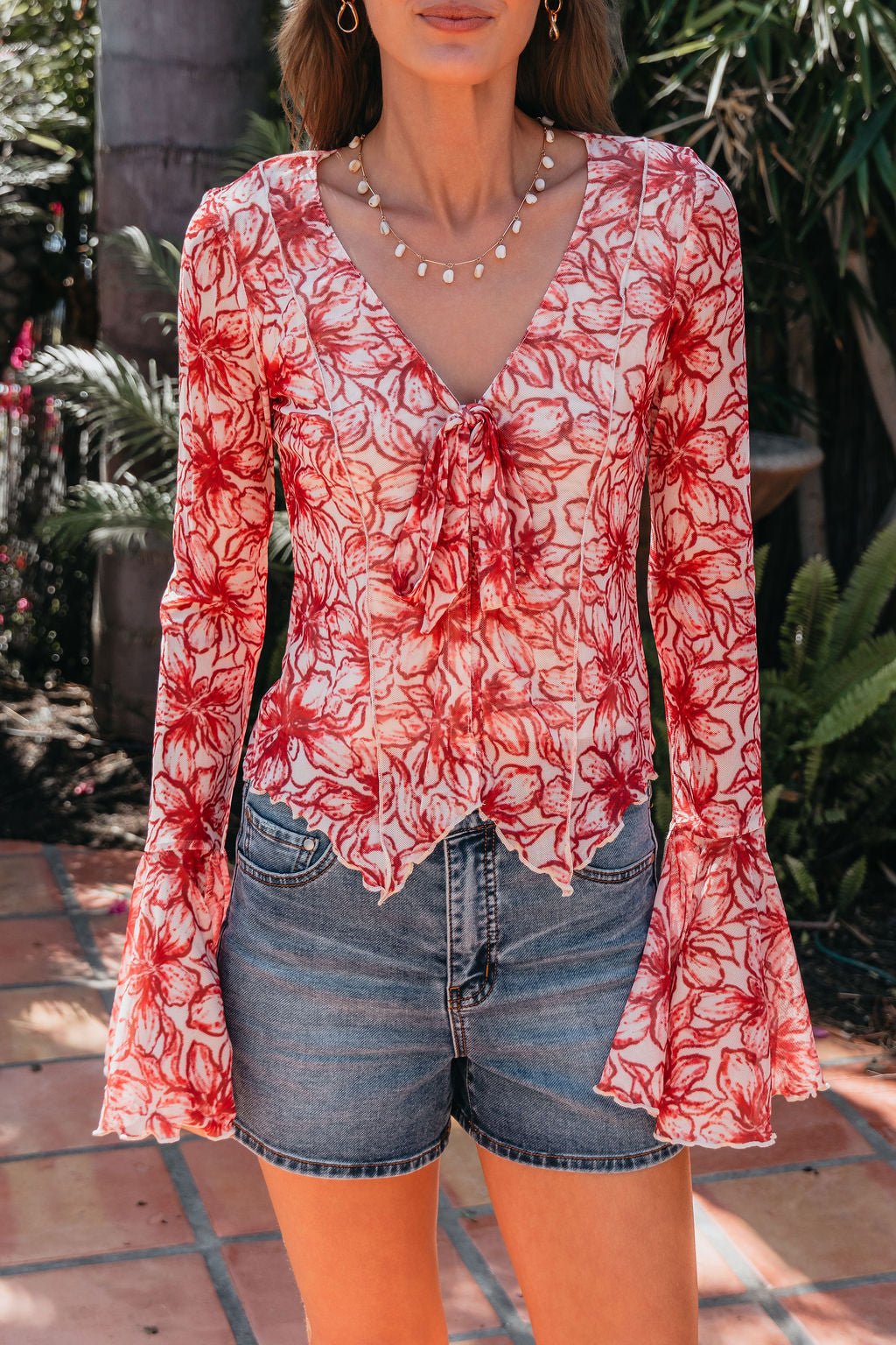 A woman wears the Red Floral Print Bell Sleeve Mesh Top - FINAL SALE with blue denim shorts, standing near plants outdoors.