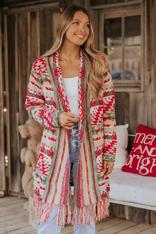 A woman smiles on a porch in the Red & Green Aztec Striped Boho Fringe Cardigan—perfect for festive Christmas layering.