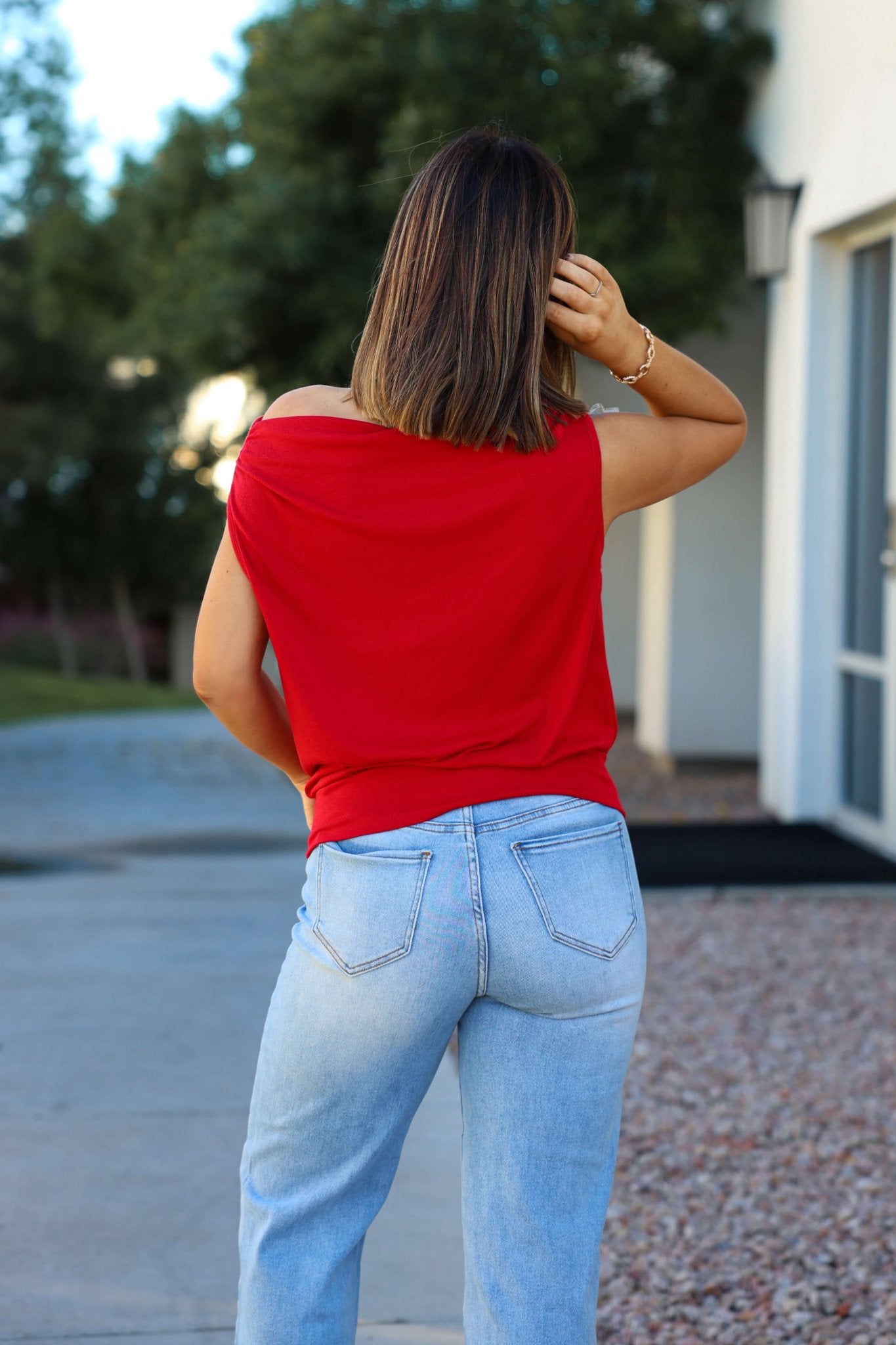 A woman in a Red Jersey Knit Off The Shoulder Top and light blue jeans stands outdoors, facing away, hand in her hair.