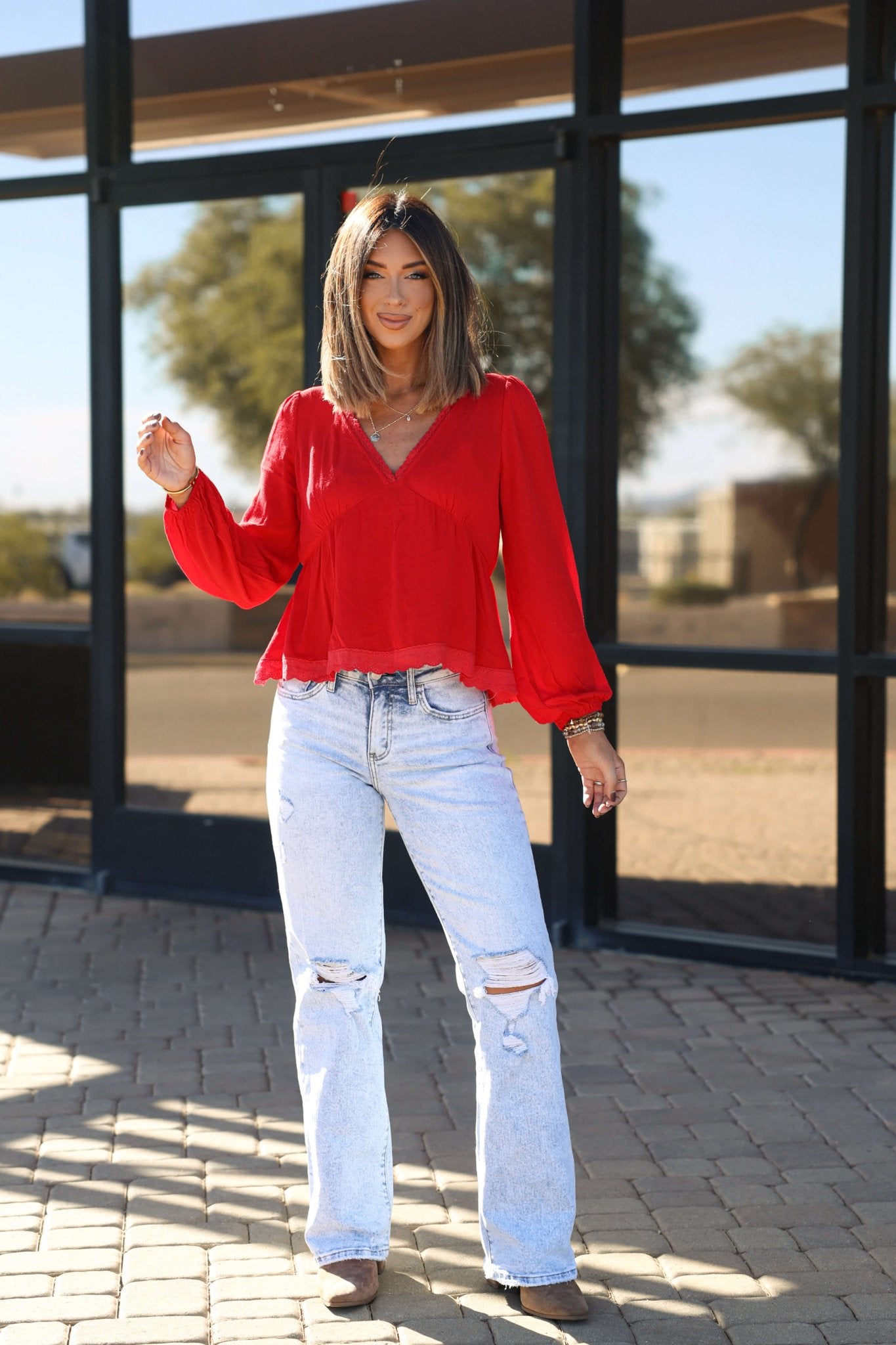 Woman in a Red Lace Trim Babydoll Top - FINAL SALE and ripped jeans smiles outdoors, showcasing a chic, feminine fall style.