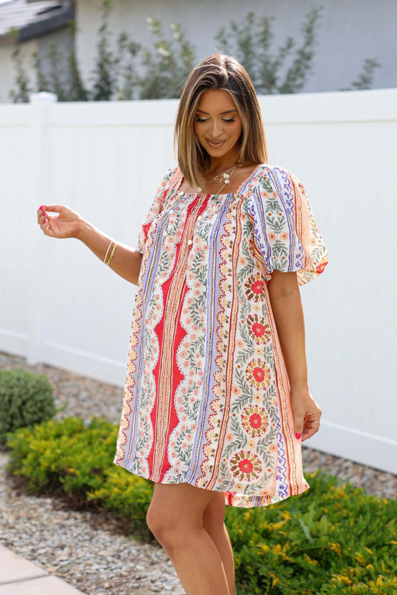 Woman wears the Red Mix Print Puff Sleeve Mini Dress - FINAL SALE outdoors near green bushes and a white fence.