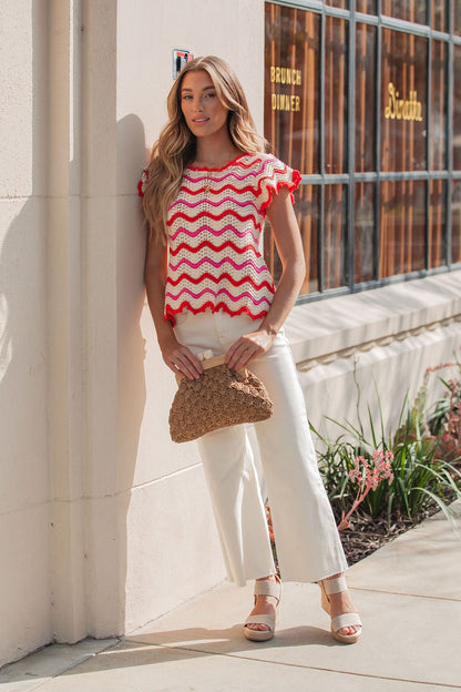 Woman in a Red Multi Chevron Knit Sweater, white pants, and sandals holds a woven bag by a restaurant window.