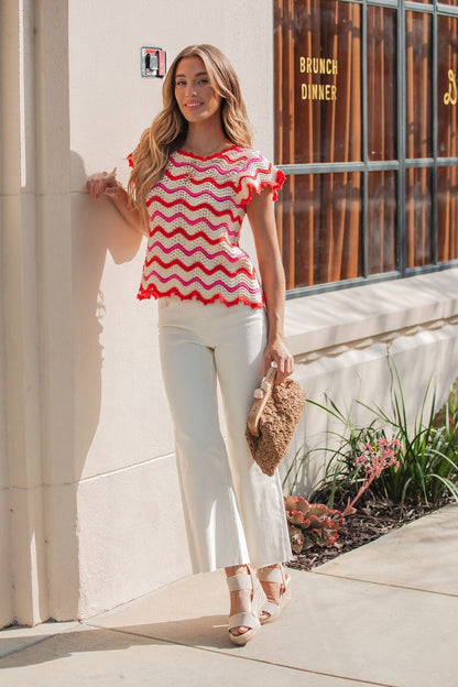 A woman in a Red Multi Chevron Knit Sweater, white pants, and sandals stands outside, holding a woven clutch.