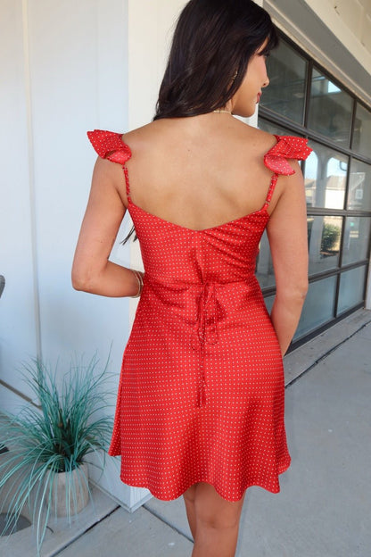 Woman wearing a Red Polka Dot Satin Mini Dress with tied straps and open back stands outside by a building with large windows.