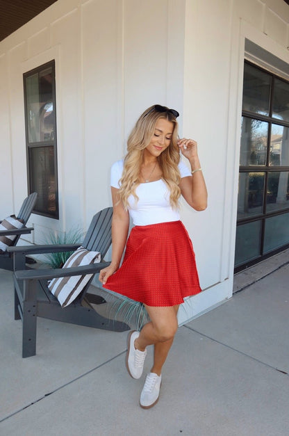 Woman in a white top and Red Polka Dot Satin Mini Skirt poses outdoors by gray chairs and large windows.