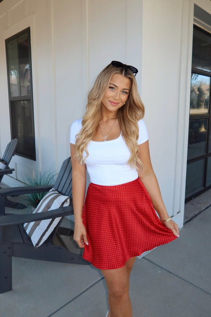 A smiling woman in a white top and the Red Polka Dot Satin Mini Skirt stands outside near gray patio chairs.