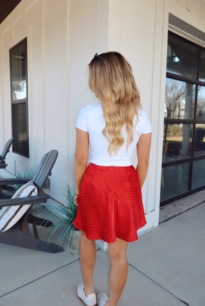 A woman in a Red Polka Dot Satin Mini Skirt and white top stands outside a house with large windows, facing away.