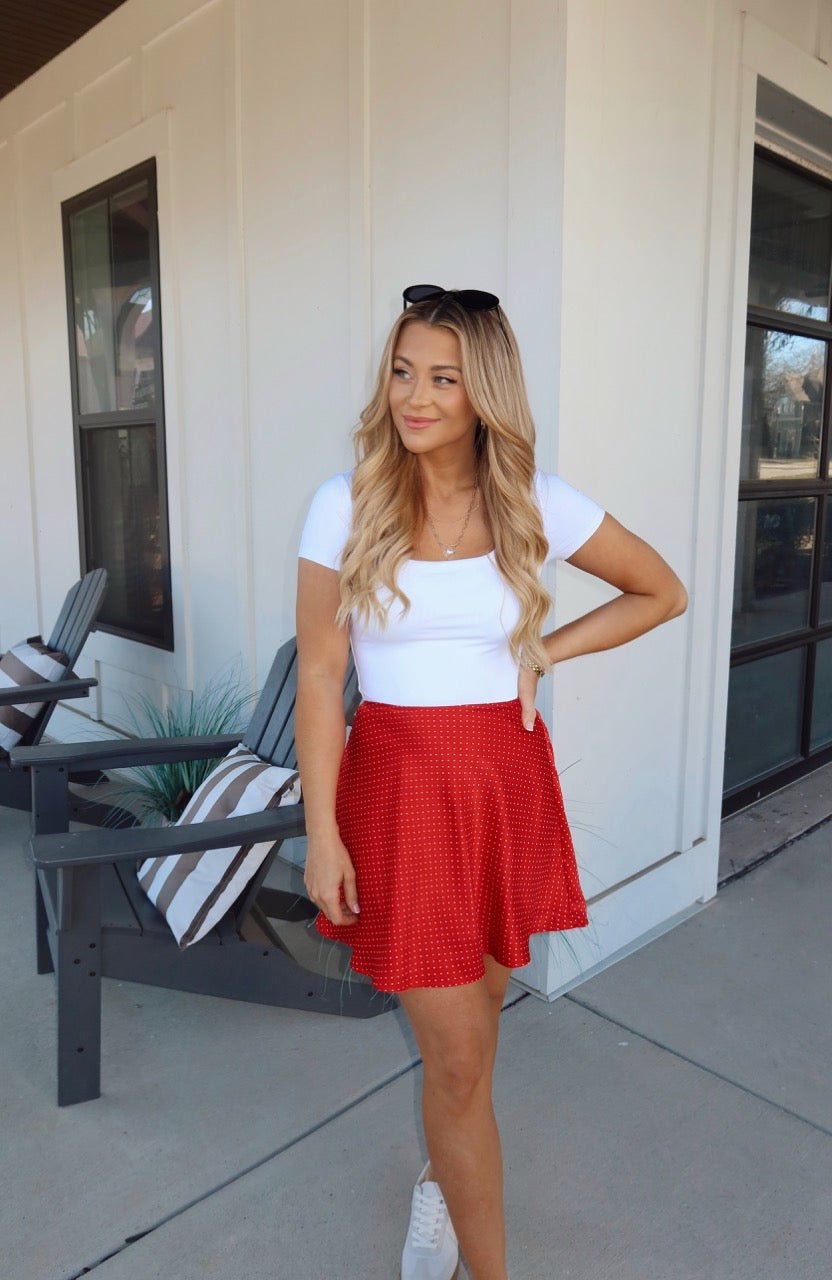 Smiling woman with long blonde hair wears a Red Polka Dot Satin Mini Skirt and white top, posing outdoors with sunglasses on her head.