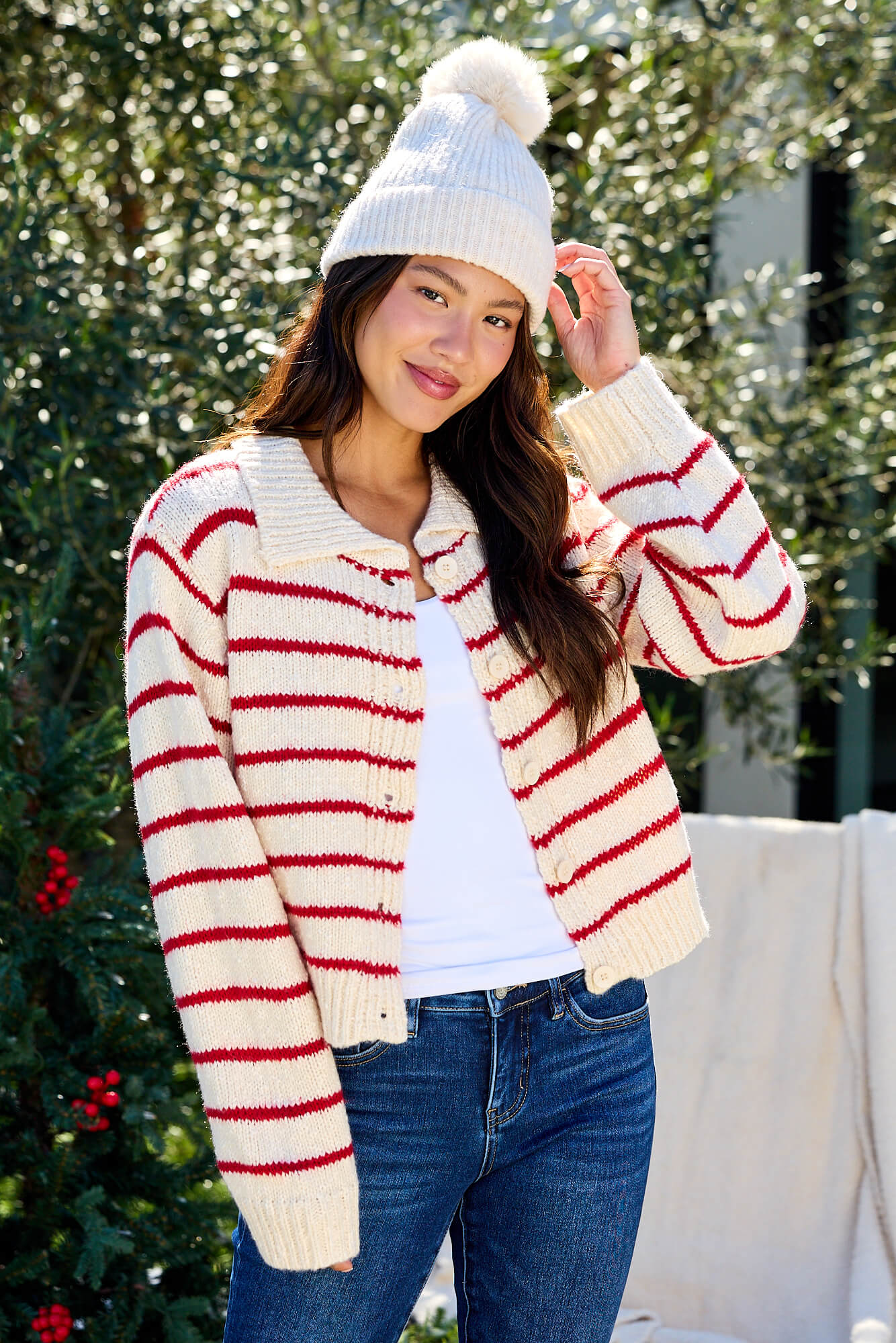 A woman smiles outdoors in jeans and a Red Striped Button Down Knit Cardigan - FINAL SALE, standing near green foliage.