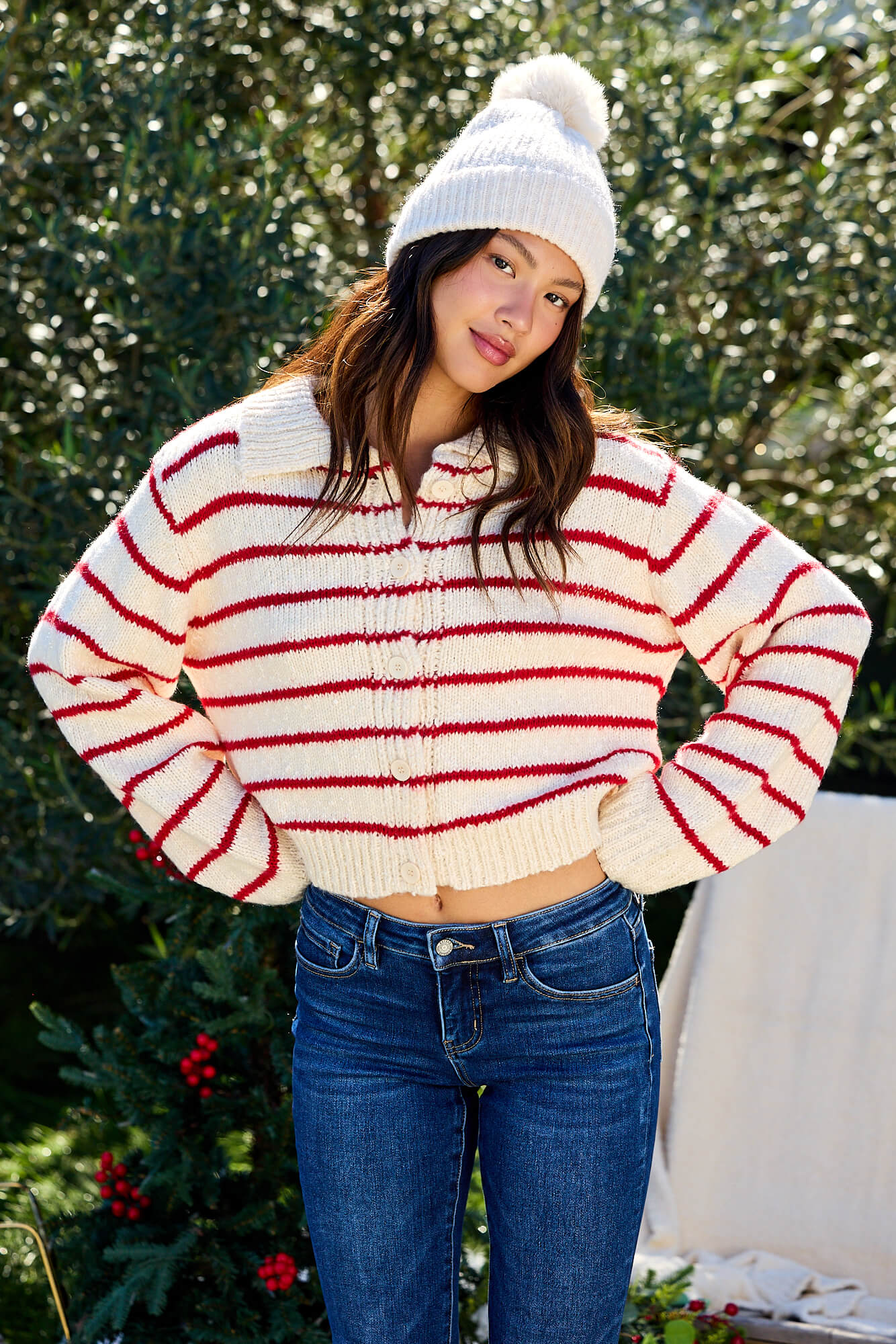 A woman wears the Red Striped Button Down Knit Cardigan - FINAL SALE and a white beanie, posing outdoors with a slight smile.