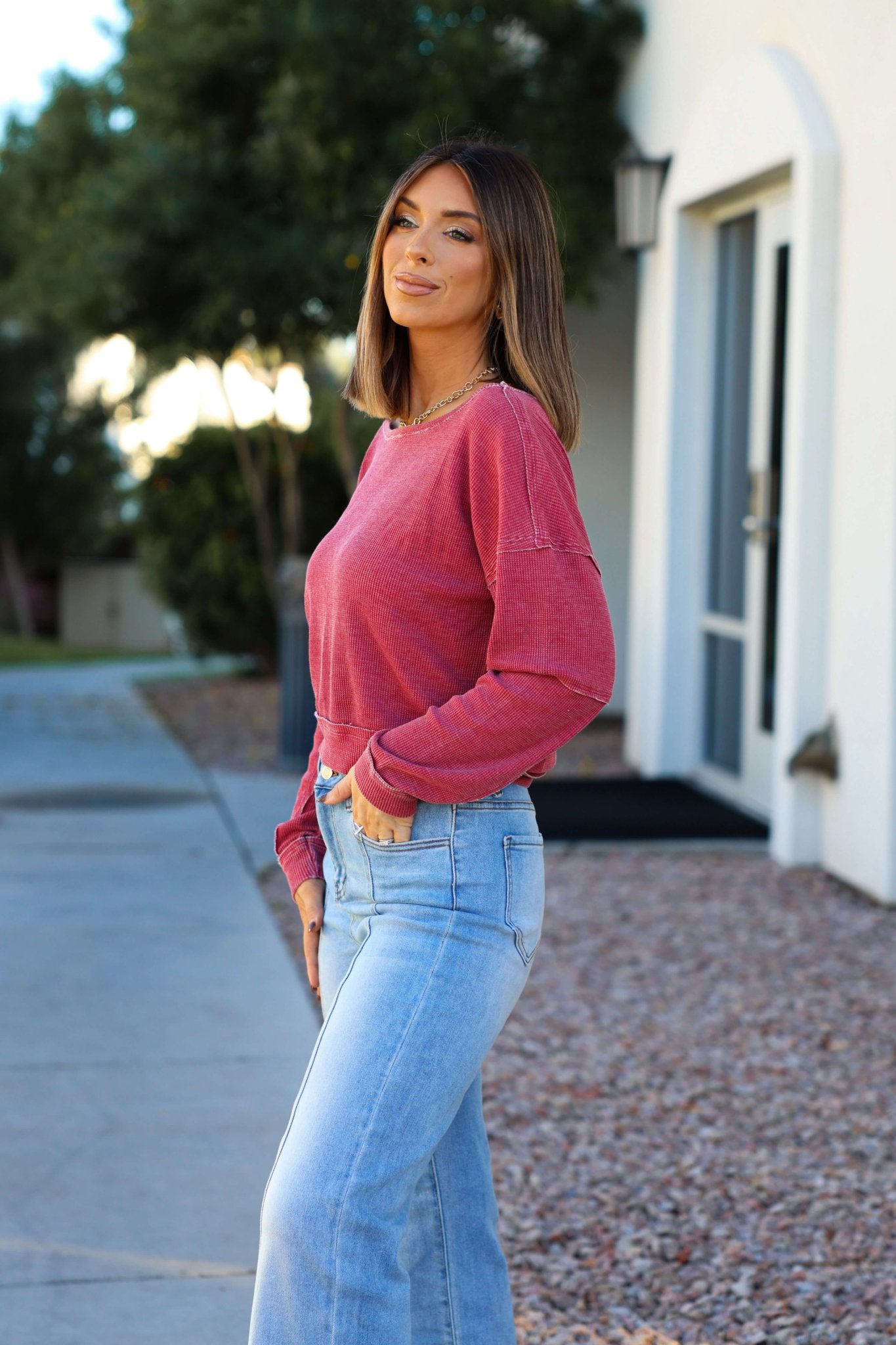 Woman with shoulder-length brown hair in a Red Waffle Knit Boatneck Pullover and light blue jeans, standing outside by a white building.