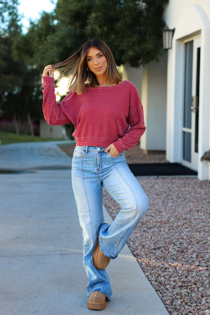 Woman poses outdoors by a modern building in a Red Waffle Knit Boatneck Pullover, blue jeans, and tan shoes, hand in her hair.