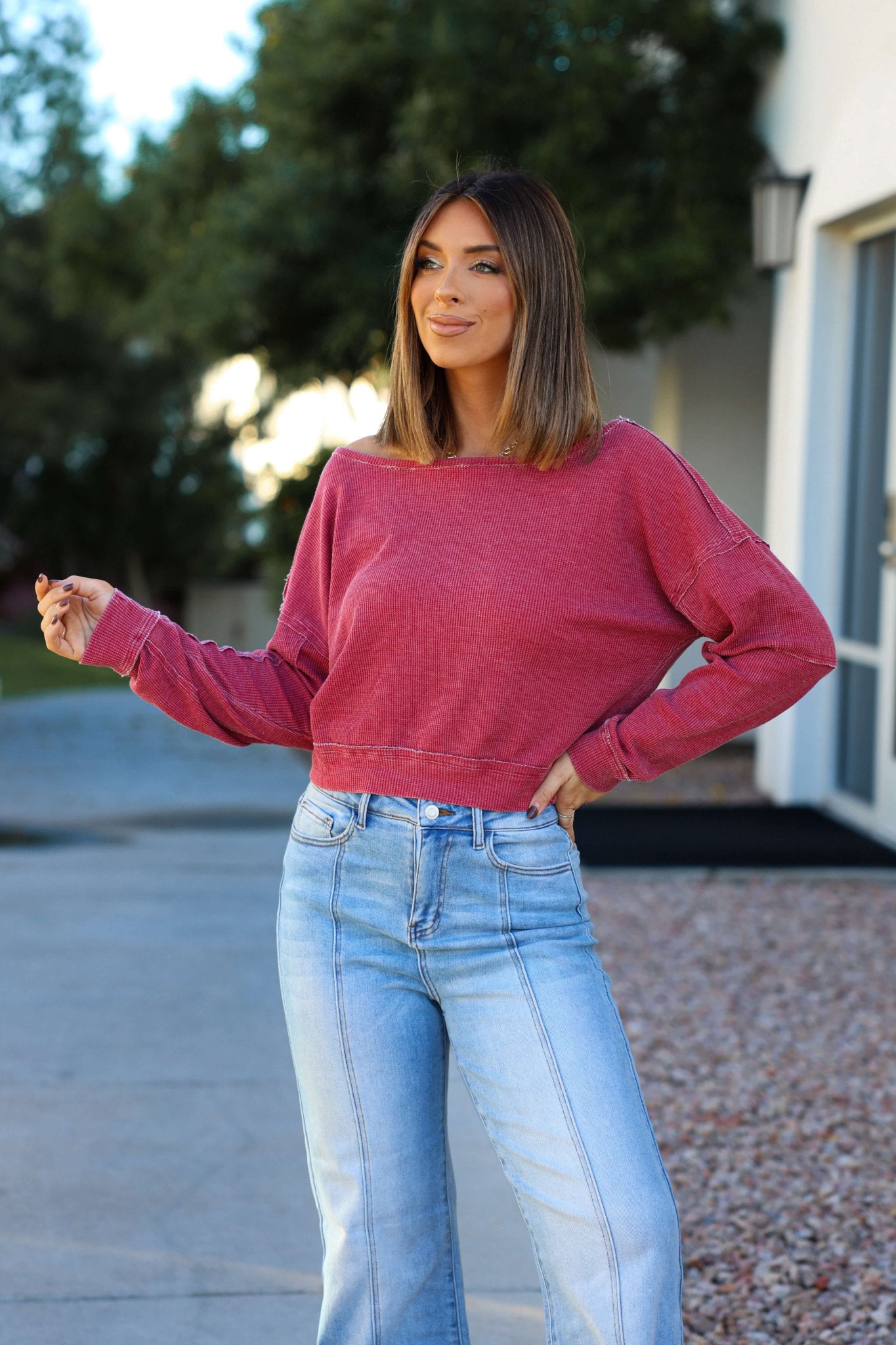 Woman outdoors in a Red Waffle Knit Boatneck Pullover and light blue jeans, with trees and a building in the background.