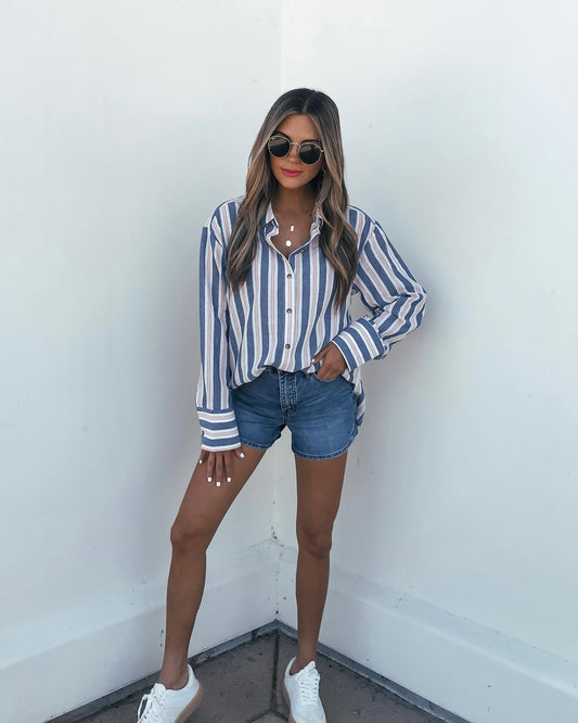Woman wears the Red White & Blue Striped Button Down Shirt with denim shorts and sneakers, standing by a white wall.