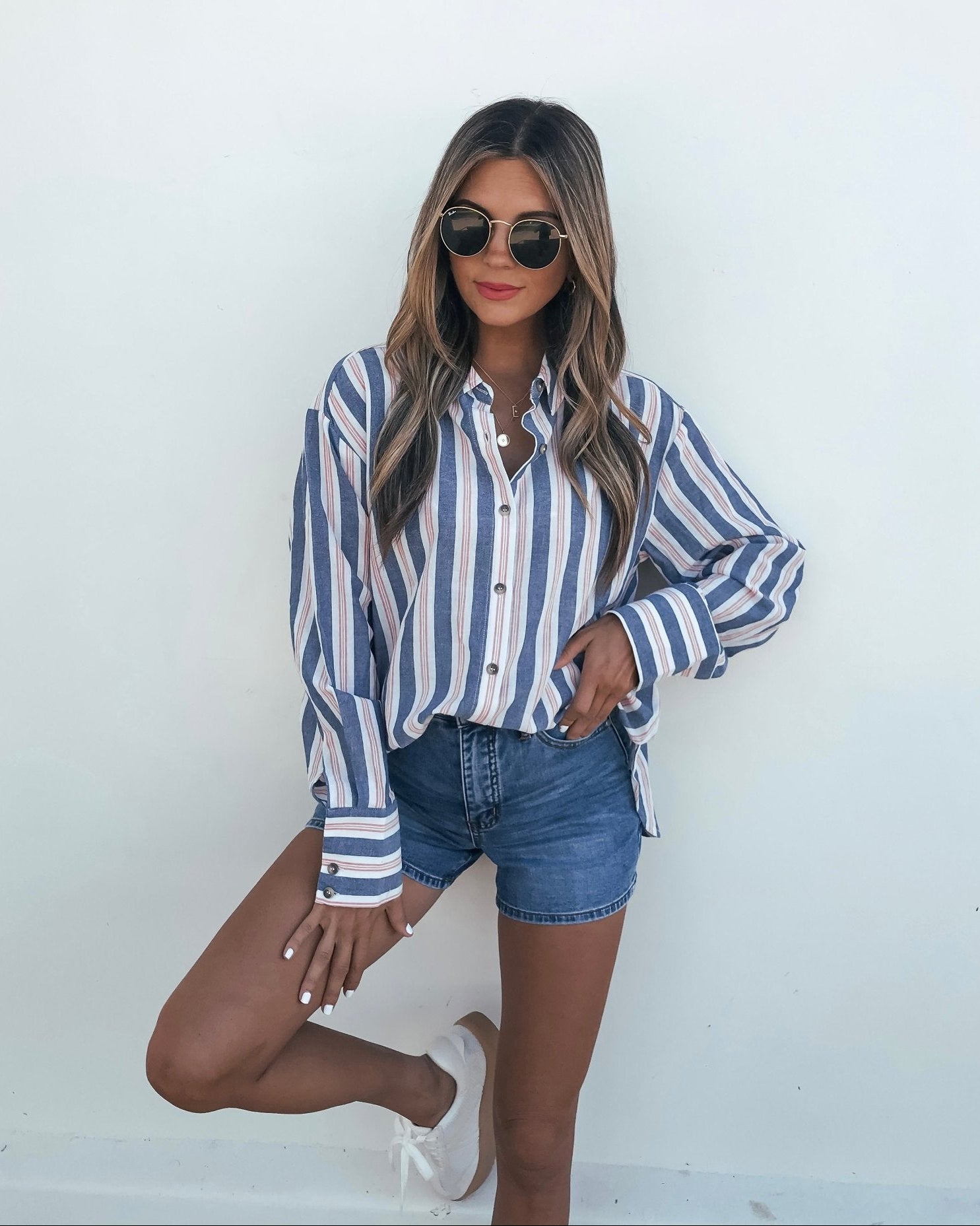 Woman wearing the Red White & Blue Striped Button Down Shirt - FINAL SALE, denim shorts, and sneakers against a white wall.